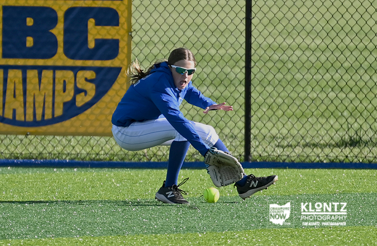 Young female softball player in a blue hoodie and white pants fields a ground ball with her glove near a chain-link fence behind her.