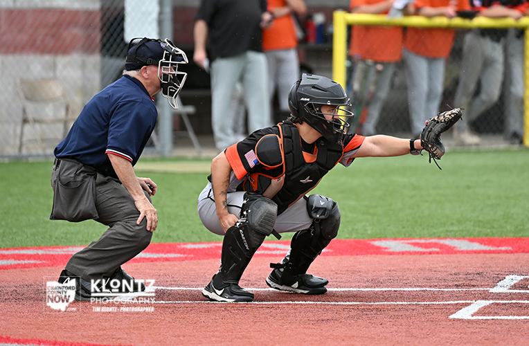 Youth baseball catcher in black and orange protective gear crouches at home plate with glove extended, umpire behind ready to call the play.