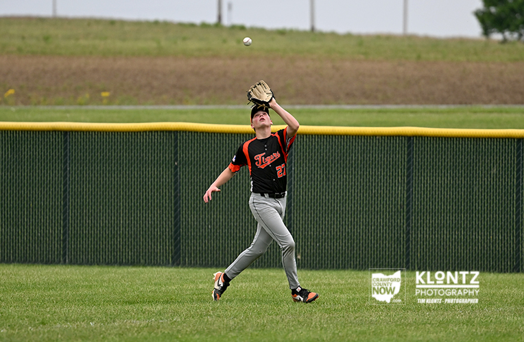 Young baseball player in a black and orange Texas uniform leaps with glove raised to catch a ball on a green outfield, yellow fence behind him.