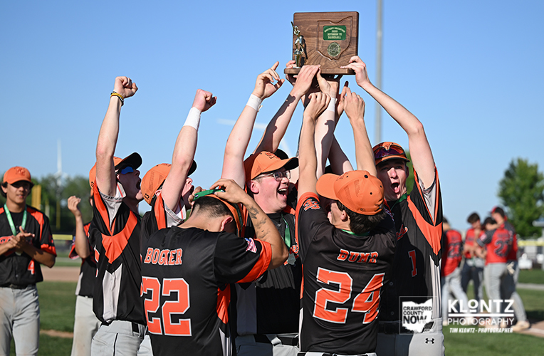 Baseball teammates in orange and black celebrate on a sunny field as they lift a wooden trophy overhead.