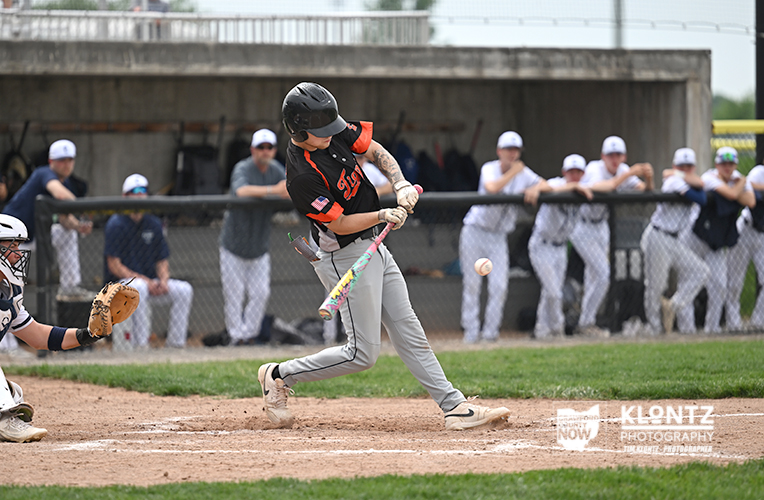Baseball batter in black and orange swings at a pitch, ball just before the bat, with catcher and dugout spectators behind the fence.