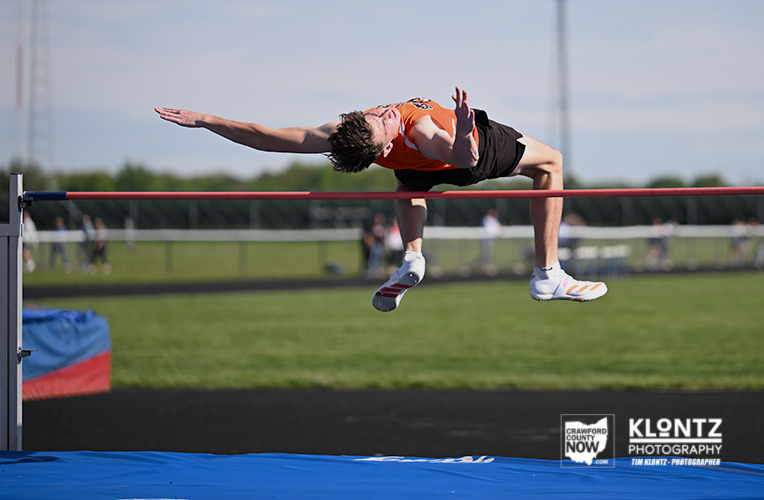 Male high jumper in an orange shirt clears the bar over the landing mat on a sunny track field.