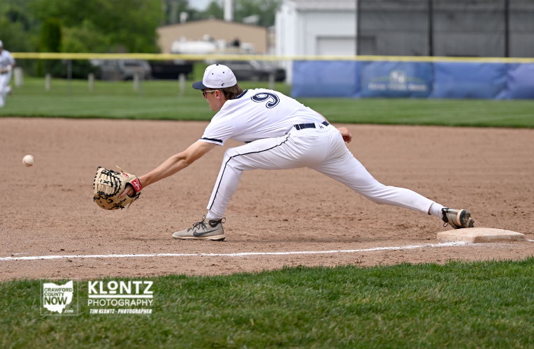 Baseball infielder in white uniform lunges to field a ground ball, glove reaching toward the ball near a base on the dirt infield.