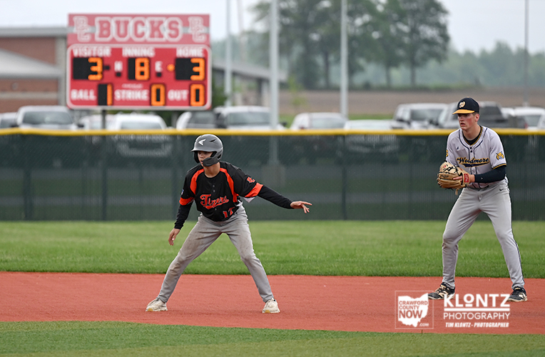Two baseball players stand on a dirt infield: one in a black-and-orange uniform bending as if to sprint, the other in a gray uniform with a glove watching from nearby, with a scoreboard and fence in the background.