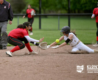Softball game action: a red-clad fielder dives to tag a sliding white-uniform runner at a base, both reaching for the ball near the bag on a dirt infield.