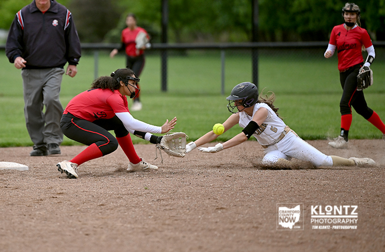 Softball game action: a red-clad fielder dives to tag a sliding white-uniform runner at a base, both reaching for the ball near the bag on a dirt infield.