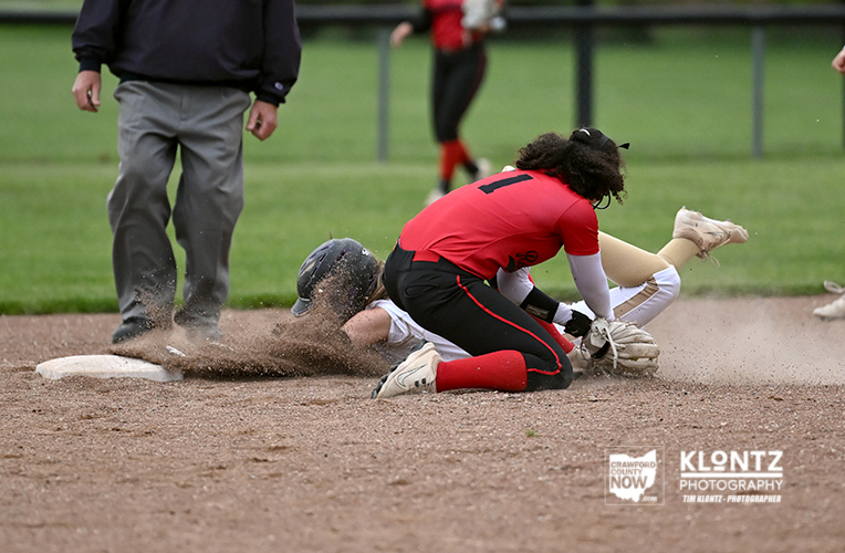 Baseball player in a red jersey slides into a base as a fielder on the ground reaches for the ball; dirt flies on the infield.