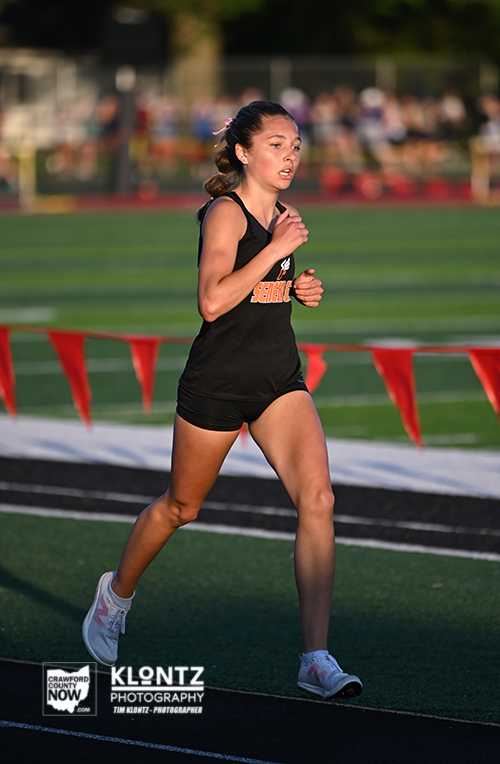 Female runner in a black uniform mid-stride on a track during a race, with red banners in the background.
