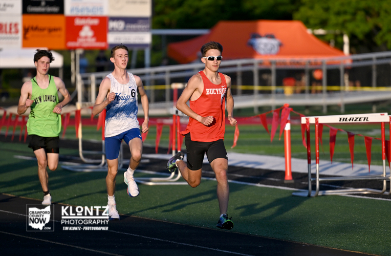 Three male sprinters run at a outdoor track meet; the lead runner wears a red singlet and sunglasses, with two others in white/blue and neon green behind him, lanes lined with barriers and sponsor banners.
