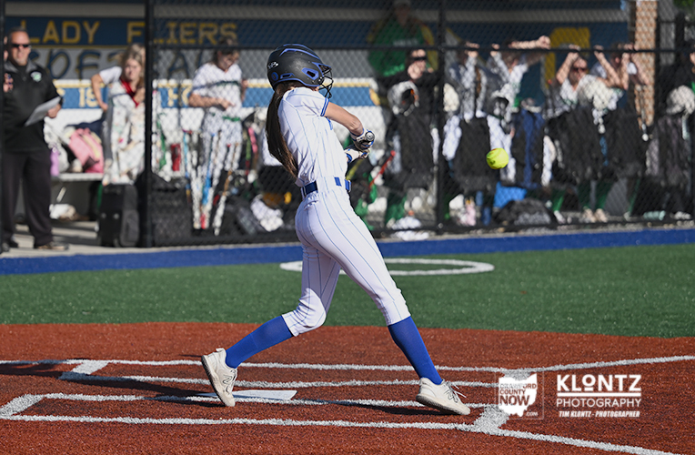 Female softball player in a white and blue uniform swings at a pitch on a fenced field with spectators in the background.