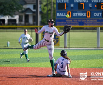 Baseball scene: pitcher in mid-throw on the mound as a baserunner slides into a base, with a scoreboard visible in the blue outfield behind them.