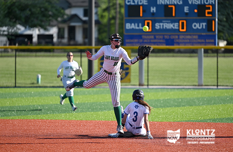 Baseball scene: pitcher in mid-throw on the mound as a baserunner slides into a base, with a scoreboard visible in the blue outfield behind them.