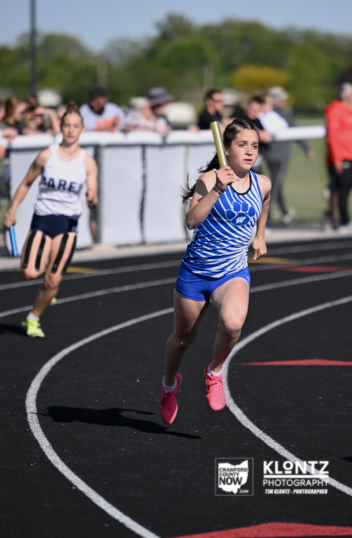 Girl in a blue relay uniform runs with a baton on a curved track as spectators watch from the background.