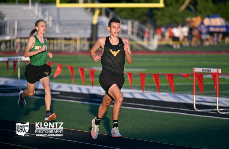 Two high school or college sprinters race on a track, one in a black uniform with a gold logo and the other in green, near the finish line with red flags nearby.
