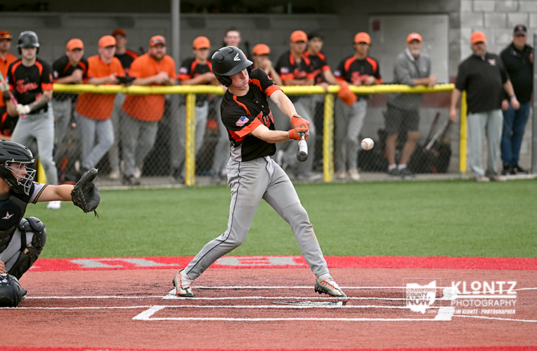 Baseball batter in black and orange swings at a pitch as the catcher crouches behind home plate and a crowd watches from the stands behind a fence.