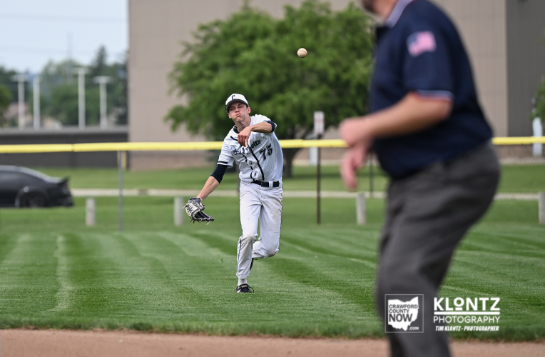 Baseball pitcher in gray uniform throws a pitch on a grassy field with an umpire blurred in the foreground.