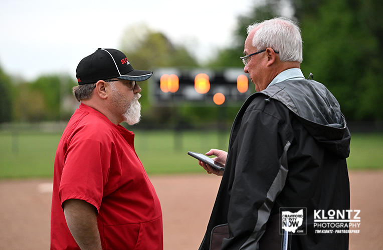 Two older men stand on a baseball field having a chat; one in a red shirt and cap, the other in a dark jacket holding a phone.