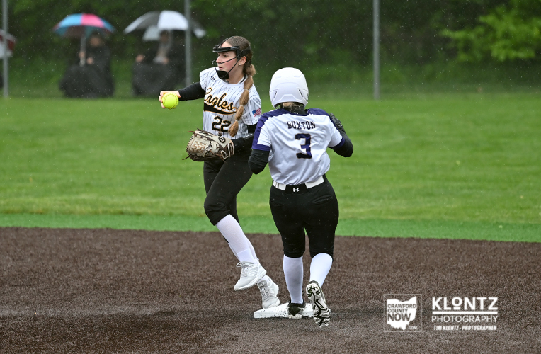 Softball action in rain: fielder with glove and ball near a baserunner on a wet infield with spectators under umbrellas in the background.