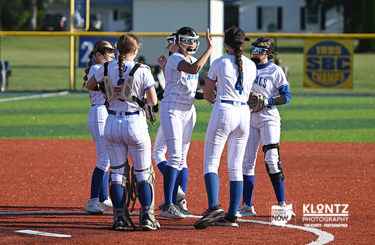 Girls softball team huddling on a red infield, high-fiving after a play during a game.