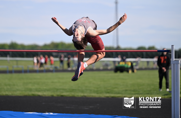 Athlete clears a high jump bar in mid-air during track and field competition, grass field and spectators in the background.
