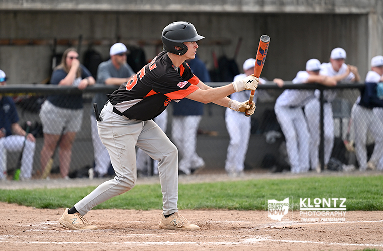 Baseball batter in a black-orange uniform swings at a pitch, with spectators behind a chain-link fence watching.