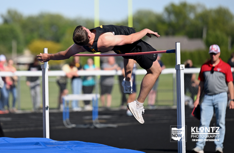 Male high jumper clears the bar mid-air during a track and field event, with onlookers and officials in the background.