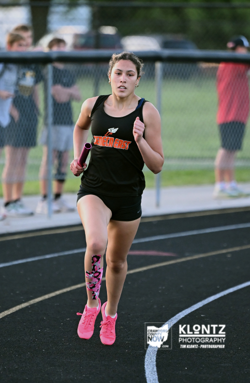 Female sprinter in a black uniform running on a track with a pink baton, spectators behind a chain-link fence; pink shoes and a taped leg highlight her motion.