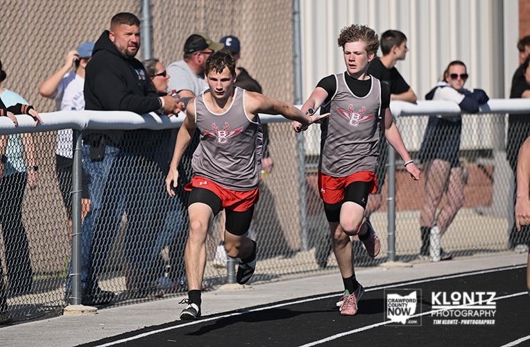 Two male relay runners in gray singlets and red shorts sprint on a track; one hands off a baton in a relay.