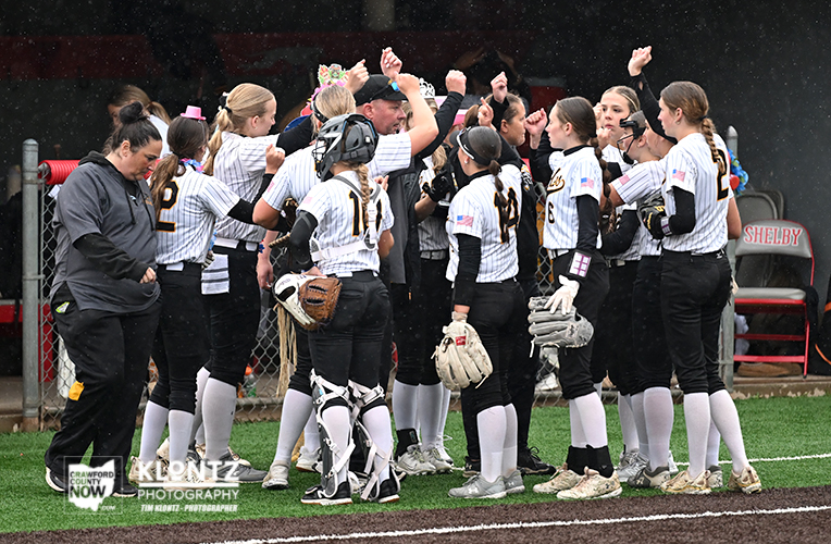 Girls softball team in white striped uniforms huddle on the field, listening to a coach at center.00