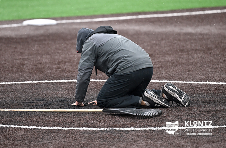Person kneeling on a baseball infield, measuring a line with a tape measure.