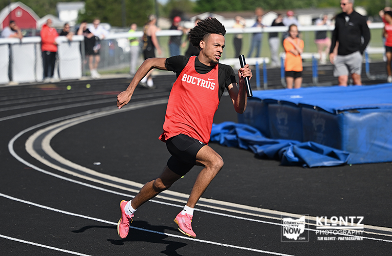 Male sprinter in a red Buckyros jersey runs on a track holding a baton during a relay race with spectators in the background.