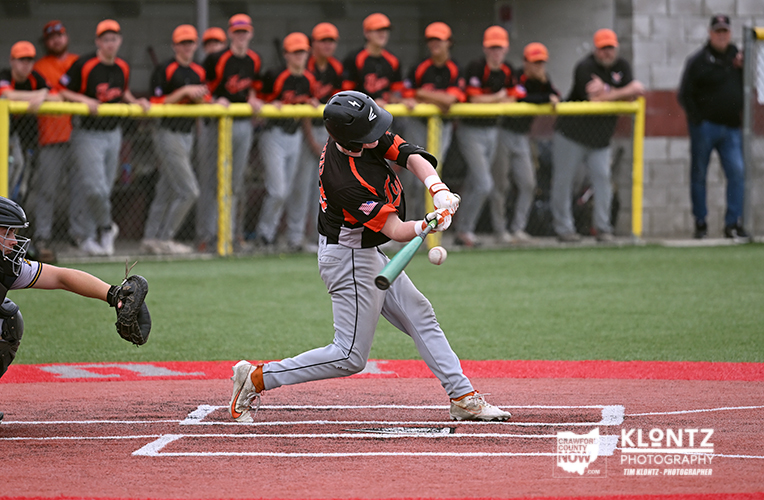 Baseball batter in black and orange swings at a pitch with the catcher behind him and spectators in the background.