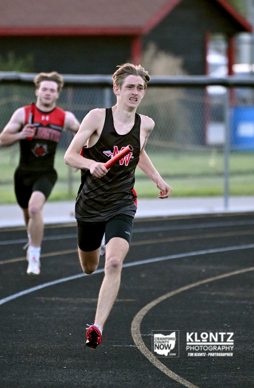 Two male relay runners sprint on an outdoor track; the front runner in a black jersey holds a red baton, focused on the race.