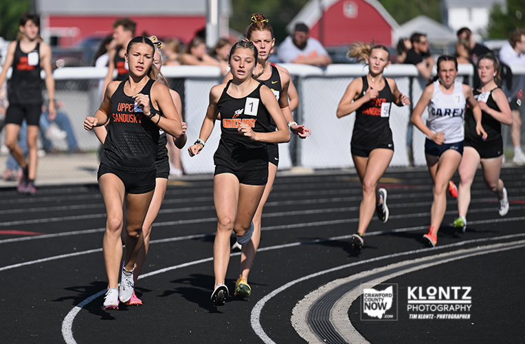 Girls' track sprint race on an outdoor track; front runners wear black uniforms with bib numbers; crowd and fence in the background.