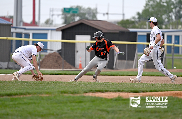 Baserunner in black and orange slides into a base as two players in white uniforms reach for the ball on the infield dirt.