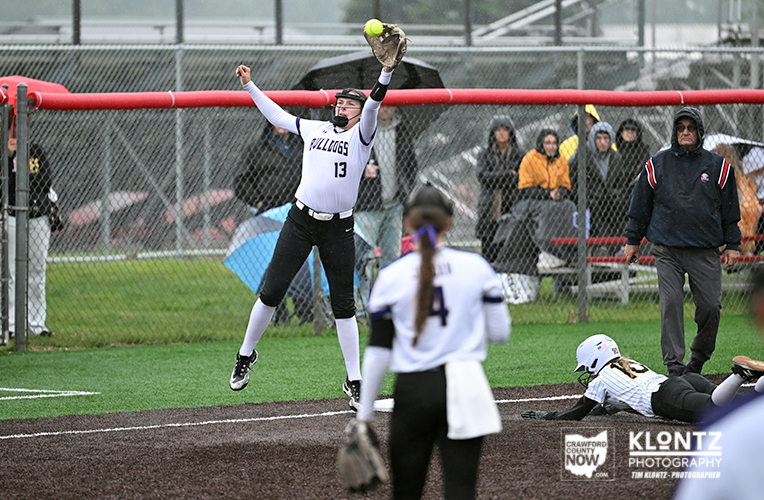 Softball pitcher in white uniform (#13) throws a pitch while leaping near home plate, with a catcher on the ground and spectators in the dugout area behind a chain-link fence.
