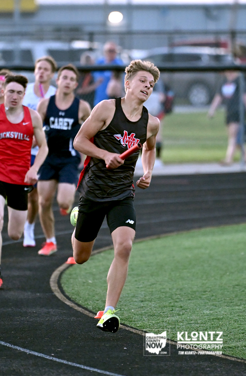 Young male sprinter in black leading a relay on an outdoor track, baton in hand, with teammates behind him.