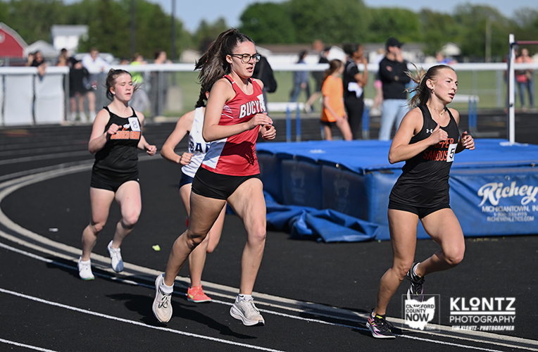 Four female track runners sprint on a black outdoor track during a race, with spectators and blue safety mats in the background.