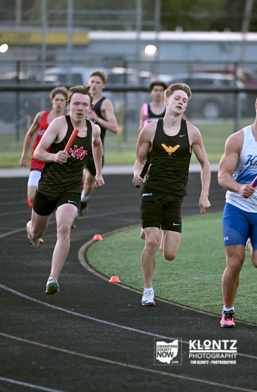 Young male sprinters racing on an outdoor track during a relay or sprint event, wearing black uniforms with colorful logos, mid-stride toward the finish line.