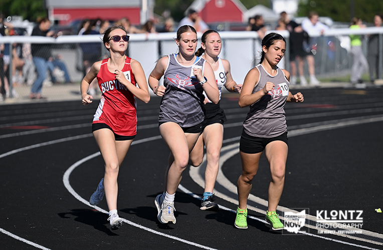 Four female sprinters race on an outdoor track, wearing colorful uniforms with numbers, focused on the finish line.