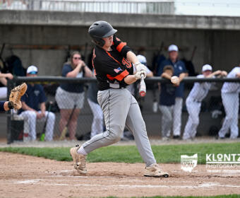 Baseball batter in a black and orange uniform mid-swing, with a ball just off the bat and the catcher reaching from the left.