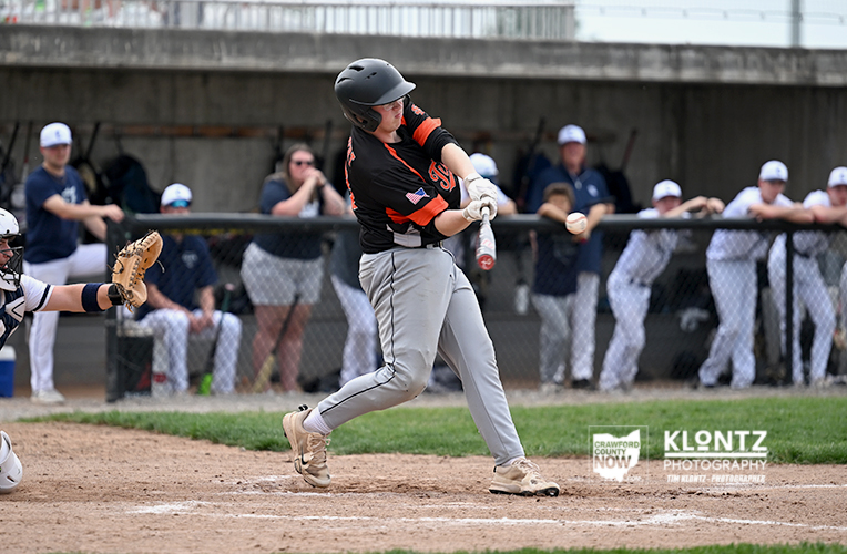 Baseball batter in a black and orange uniform mid-swing, with a ball just off the bat and the catcher reaching from the left.