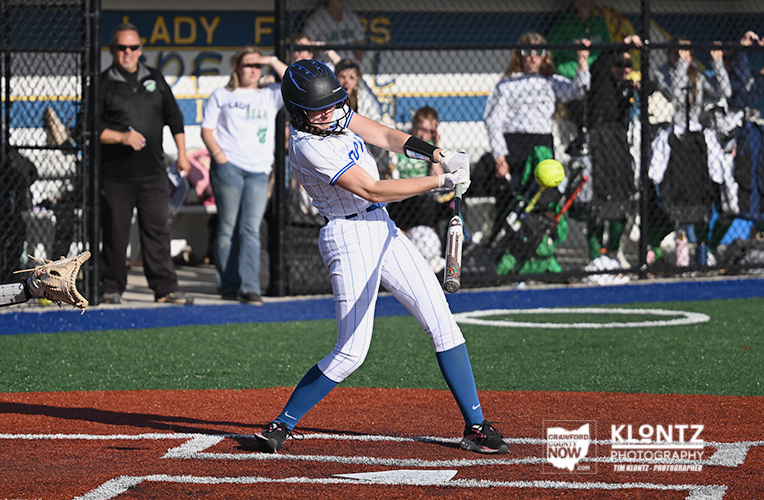 Female softball batter in white pinstripes swings at a pitch on a turf field with spectators behind the fence.