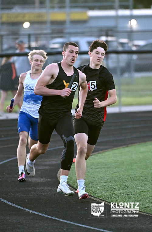 Three male sprinters race on an outdoor track; front runner in black (bib 2) followed by a runner in black (bib 5) and a blue/white-clad racer behind; motion blur and fence in background.