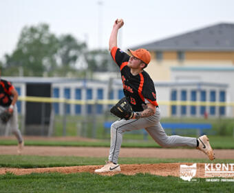 Baseball pitcher in orange and black uniform mid-throw on the mound, with teammate in the background and field goalposts visible nearby.