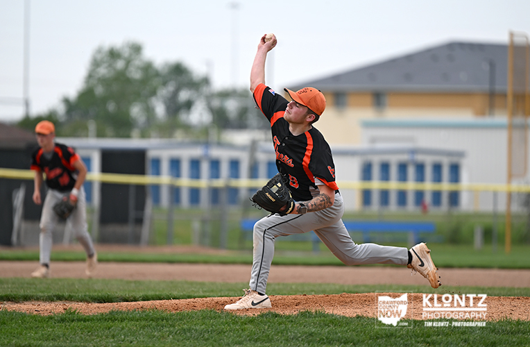 Baseball pitcher in orange and black uniform mid-throw on the mound, with teammate in the background and field goalposts visible nearby.