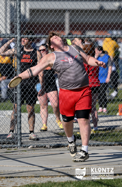 Male discus thrower in a gray sleeveless shirt and red shorts releases the discus mid-throw at an outdoor meet, with a chain-link fence and onlookers in the background.