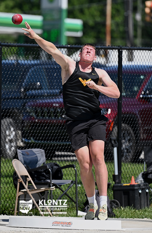 Male shot put athlete in a black sleeveless shirt and shorts releases a red shot put from a throwing circle on a sunny day, mid-throw.