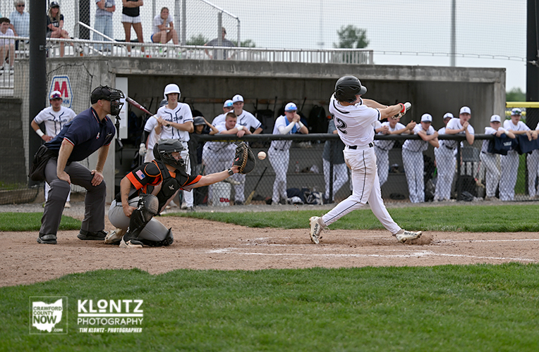 Baseball batter swings at a pitch near home plate as catcher crouches and umpire looks on. Behind them, dugout and spectators watch from the fence.
