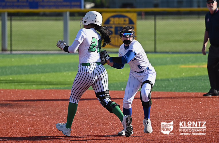 Two softball players in white uniforms compete for the ball at a base: the baserunner (number 20) with green socks is advancing while the fielder, wearing blue sleeves and a helmet, reaches to tag.